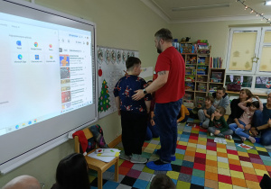 Dzieci z rodzicami siedzą w półkolu, a chłopiec szuka podpisu do swojego obrazka z pomocą taty.