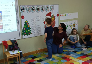 Dzieci z rodzicami siedzą w półkolu, a chłopiec szuka podpisu do swojego obrazka z pomocą nauczycielki.