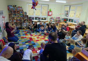Dzieci z rodzicami siedzą w półkolu na dywanie, a nauczycielka opowiada o czym będą zajęcia.