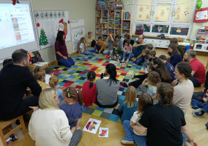 Dzieci z rodzicami siedzą w półkolu na dywanie, chłopiec nazywa przedmiot znajdujący się na jego puzzlach.