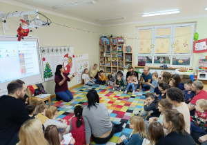 Dzieci z rodzicami siedzą w półkolu na dywanie, a nauczycielka mówi rymowankę na powitalnie.