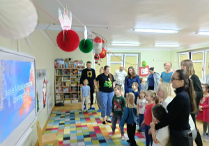 Rodzice z dziećmi stoją i wspólnie śpiewają piosenkę wyświetlaną na tablicy multimedialnej podczas zabawy karaoke.