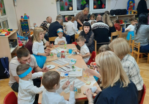 Rodzice wraz z dziećmi siedzą przy stolikach i ozdabiają szklane lampiony.