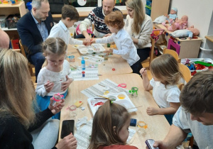 Rodzice wraz z dziećmi siedzą przy stolikach i ozdabiają szklane lampiony.