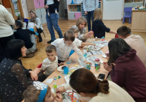 Rodzice wraz z dziećmi siedzą przy stolikach i ozdabiają szklane lampiony.