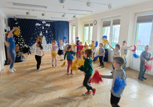 Dzieci stoją w rozsypce z kolorowymi pomponami w rękach i uczą się nowej choreografii tanecznej na zajęciach z zumby.