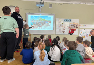 Dzieci siedzą na dywanie i oglądają prezentację przedstawiającą pracę w Muzeum Królikarni przygotowaną przez tatę kolegi w ramach projektu Bohater Tygodnia.