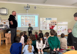 Dzieci siedzą na dywanie i oglądają prezentację przedstawiającą pracę w Muzeum Królikarni przygotowaną przez tatę kolegi w ramach projektu Bohater Tygodnia.