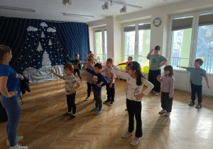Dzieci stoją w rozsypce z jedną ręką wystawioną do boku, a drugą na biodrze, uczą się nowej choreografii tanecznej na zajęciach z zumby.