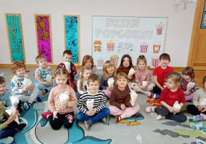 Przedszkolaki siedzą na dywanie, w dłoniach trzymają rożki wypełnione popcornem, pozują na tle dekoracji z okazji Dnia Popcornu.
