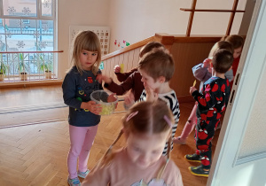 Dziewczynka trzyma w dłoniach pudełko, do którego dzieci wchodzące na salę gimanstyczną wkładają kartoniki - bilety.