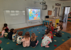 Dzieci siedzą w półkolu i słuchają piosenki projektowej wyświetlanej na tablicy interaktywnej.