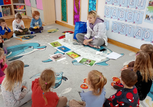 Dzieci siedzą w półkolu, przed nimi na białej ceracie rozłożone są ilustracje różnych zwierząt.