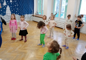 Przedszkolaki stoją w rozsypce na sali gimnastycznej, ręce trzymają na bioderkach i poruszają głowami w górę i w dół.