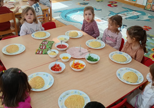 Sześć dziewczynek i dwóch chłopców siedzi wokół dwóch złączonych stolików z produktami do wykonania pizzy.