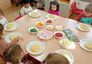 Sześć dziewczynek i dwóch chłopców siedzi wokół dwóch złączonych stolików z produktami do wykonania pizzy.