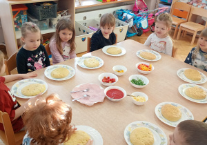 Sześć dziewczynek i czterech chłopców siedzi wokół dwóch złączonych stolików z produktami do wykonania pizzy.