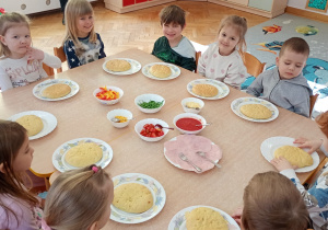 Sześć dziewczynek i czterech chłopców siedzi wokół dwóch złączonych stolików z produktami do wykonania pizzy.