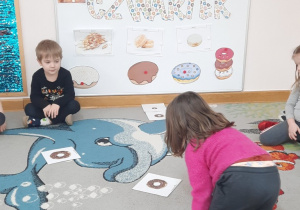 Dziewczynka szuka większego pączka od poprzedniego pośród rozłożonych na dywanie ilustracji.