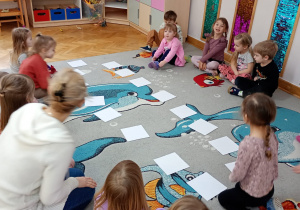 Przedszkolaki siedzą po dwóch stronach dywanu, a na środku znajdują się odwrócone do dywanu ilustracje pączków do gry w Memory.