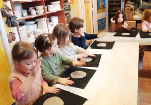 Cztery dziewczynki i chłopiec rozklepują otwartą dłonią ułożony przed nimi na ceracie kawałek gliny.