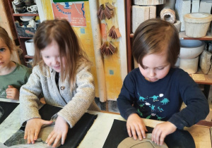 Dziewczynka za pomocą foremki wykrawa wzór serca w kawałku gliny, a siedzący obok niej chłopiec usuwa nadmiar gliny znajdujący się poza wzorem serca.