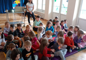 Dzieci siedzą na materacach buzią do prezentacji i słuchają prowadzącej, która stoi za nimi.