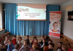 Na slajdzie widnieje pytanie: "Jak wygląda brudny ząb i co z nim wtedy zrobić?", a dzieci odwrócone do prowadzącej próbują na nie odpowiedzieć.