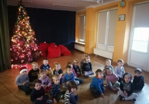 Dzieci siedzą na sali gimnastycznej i oglądają nagranie od Świętego Mikołaja.