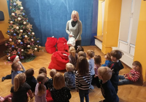 Dzieci siedzą i stoją słuchając Pani Dyrektor, która przekazuje im wiadomość od Świętego Mikołaja.