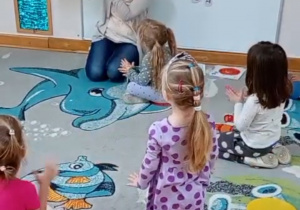 Dzieci siedzą na dywanie, oglądają pokazaną przez nauczycielkę ilustrację i klaszczą w dłonie według polecenia w książce.