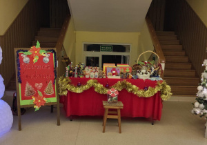Bałwanek wykonany z kubków plastikowych, plakat informujący o kiermaszu, choinka i stolik z ozdobami świątecznymi w holu przedszkola.