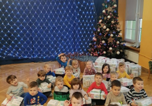 Dzieci siedzą przy choince prezentując prezenty, które zostawił im Święty Mikołaj.