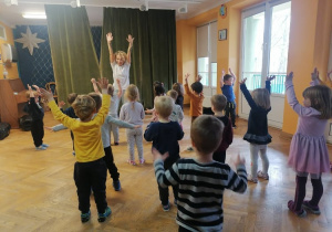 Dzieci stoją w rozsypce i wspólnie z trenerką zumby tańczą i podnoszą ręcę.
