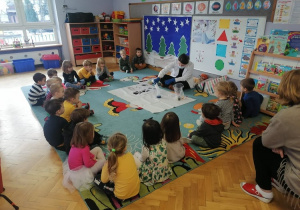 Dzieci siedzą w kole na dywanie obserwując, jak prowadzącą przeprowadza eksperyment z kolorami.