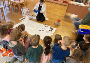 Pani instruktor pokazuje dzieciom eksperyment chemiczny na macie, a dzieci uważnie patrzą siedząc na dywanie.