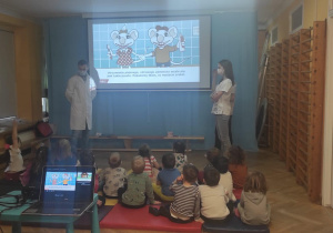 Studenci stomatologii pokazują na prezentacji zasady prawidłowej higieny jamy ustnej. Dzieci siedzą na materacach i uważnie słuchają.
