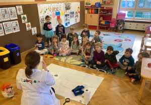Dzieci siedzą w półkole na dywanie obserwując, jak prowadzącą przeprowadza eksperyment z kolorami.