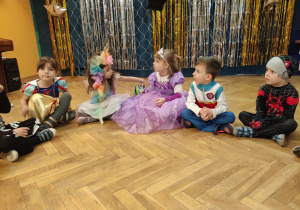 Dzieci siedzą na podłodze i słuchają poleceń nauczyciela.