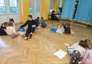 Dzieci siedzą na podłodze w trzyosobowych grupach na zajęciach z robotyki.