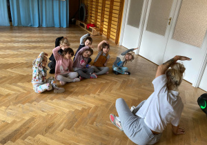 Dzieci siedzą po turecku na podłodze i patrzą na panią instruktor, która wykonuje skłon w prawo.