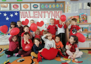 Dzieci siedzą na dywanie trzymając w dłoniach balony na tle napisu Walentynki.