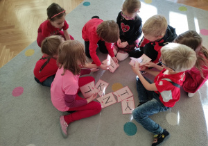 Dzieci siedzą na dywanie w małych grupkach. Z liter układają napis "Walentynki".