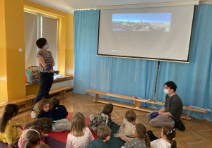 Dzieci siedzą na materacach i słuchają wykładu instruktorów na temat panoramy miast.