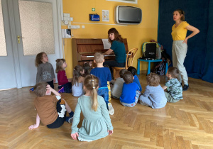Dzieci siedzą wokół pianina i śpiewają piosenkę razem z panią instruktor.