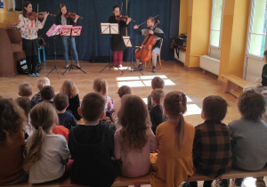 Muzycy grają na instrumentach utwór muzyki klasycznej. Dzieci siedzą na ławce i materacach słuchają muzyki.