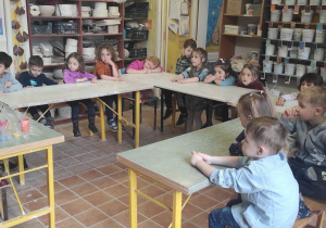 Dzieci siedzą w ławkach i słuchają wykładu pani prowadzącej warsztaty ceramiczne.