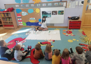 Dzieci siedzą na dywanie i obserwują jak prowadząca wykonuje doświadczenie polegające na napełnieniu balonika za pomocą drożdży.