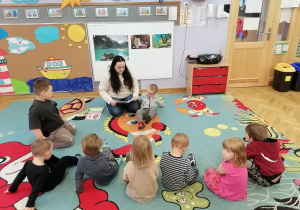 Dzieci siedzą na dywanie i słuchają jak mama Oliwiera czyta im książeczkę z bajkami.