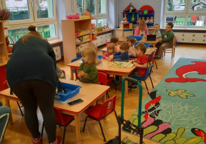 Dzieci siedzą przy stolikach i próbują skonstruować robociki z klocków Lego, przy jednym ze stolików prowadząca im w tym pomaga.